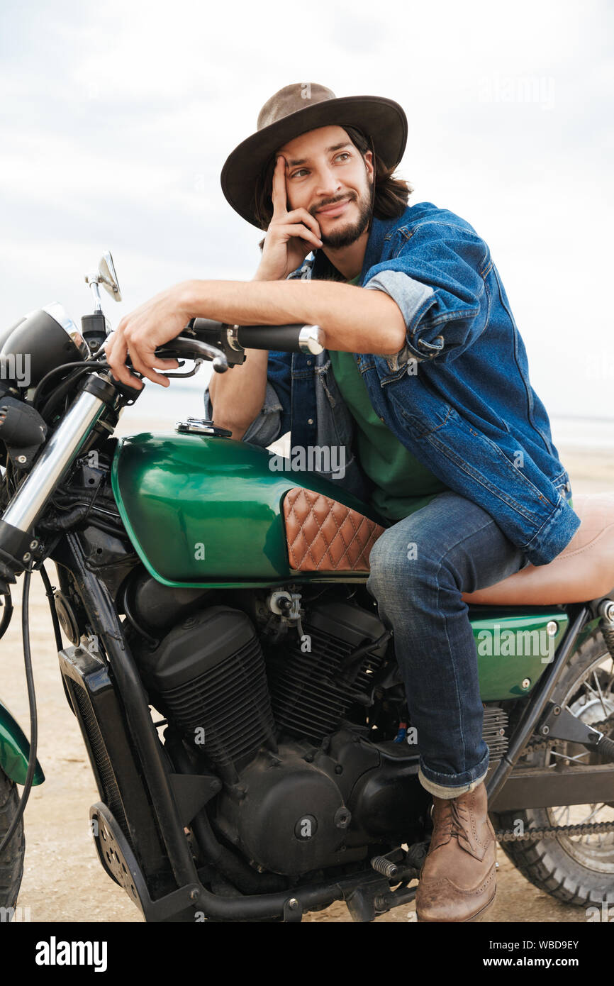 Handsome smiling young man wearing tenue décontractée assis sur une moto à la plage, de porter un chapeau Banque D'Images