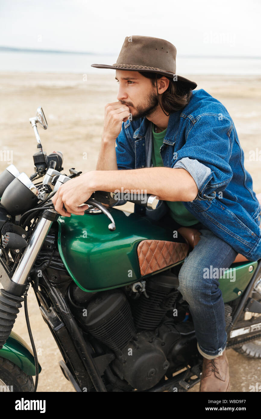 Beau jeune homme portant tenue décontractée assis sur une moto à la plage, de porter un chapeau Banque D'Images