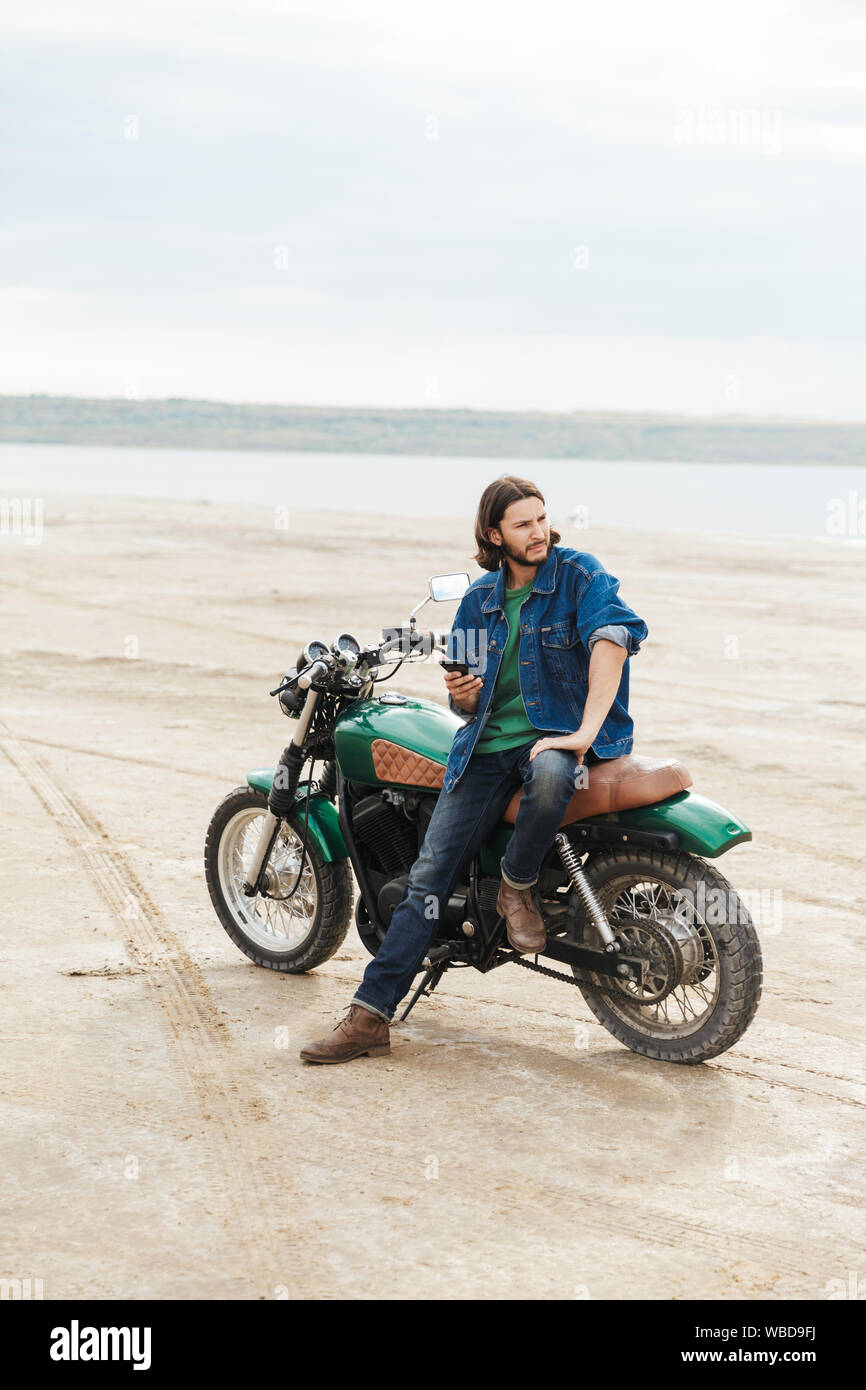 Beau jeune homme portant tenue décontractée assis sur une moto à la plage, à l'aide de mobile phone Banque D'Images