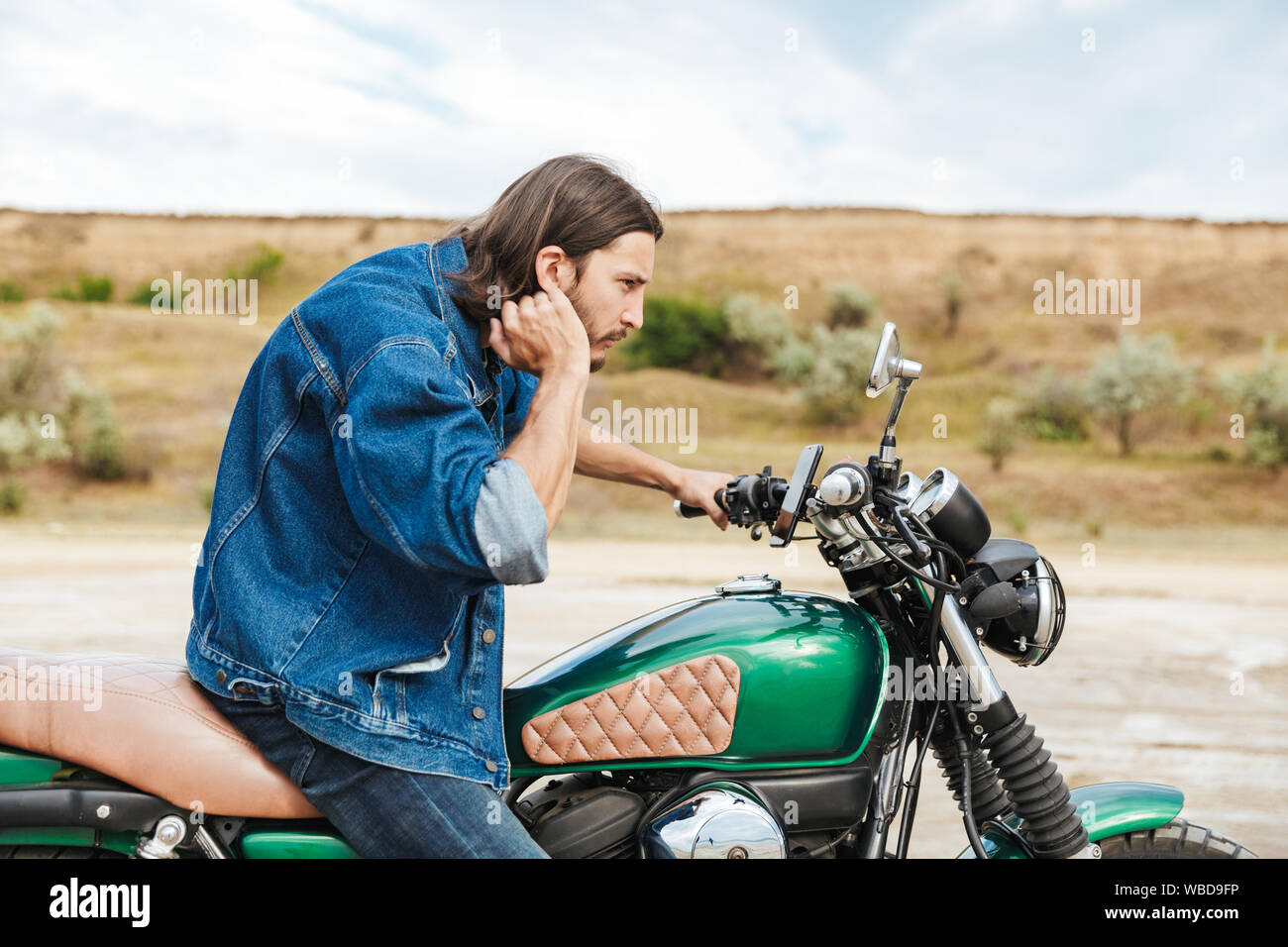Close up d'un beau jeune homme portant tenue décontractée assis sur une moto à la plage, regardant le miroir Banque D'Images