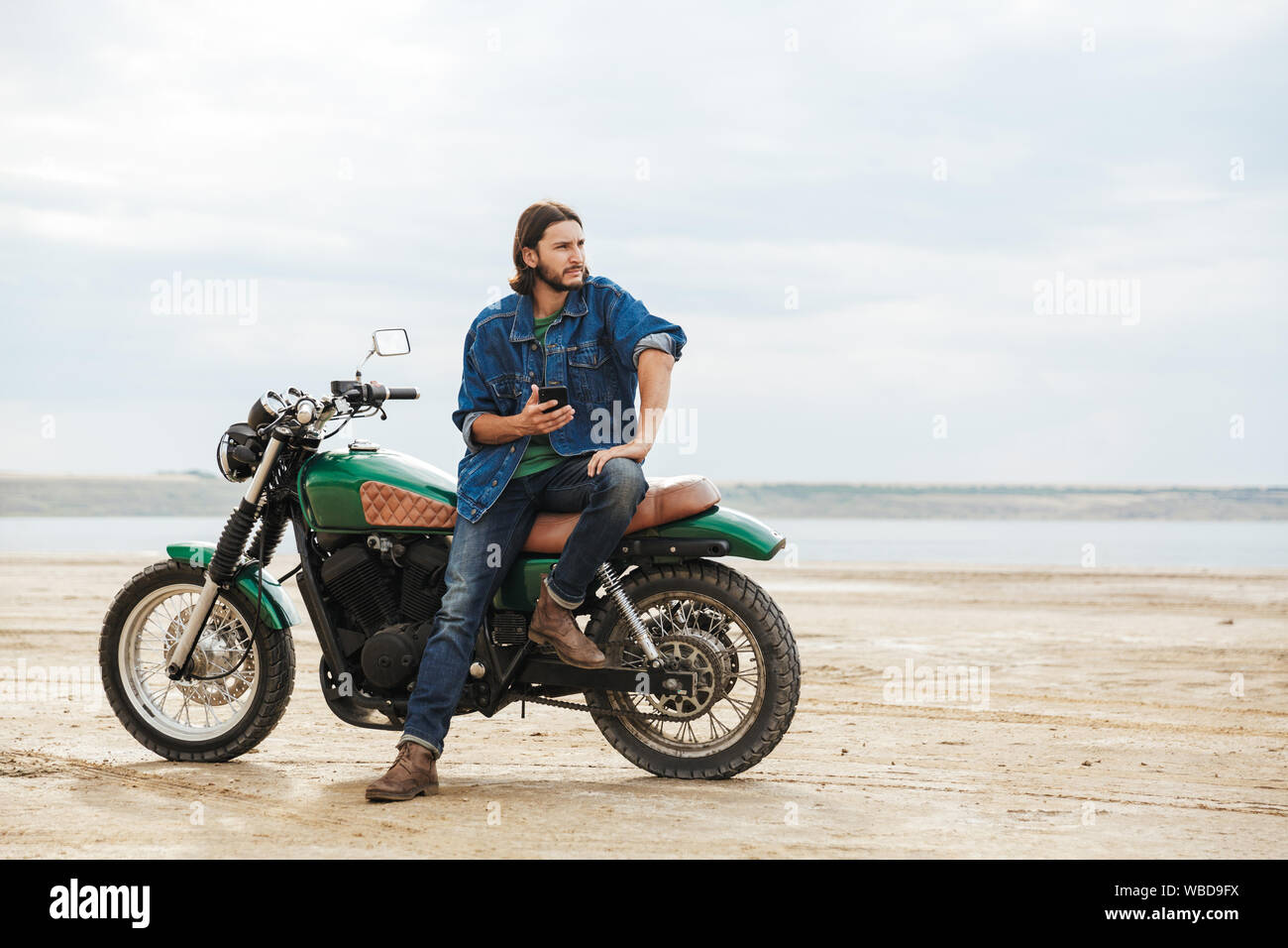 Beau jeune homme portant tenue décontractée assis sur une moto à la plage, à l'aide de mobile phone Banque D'Images