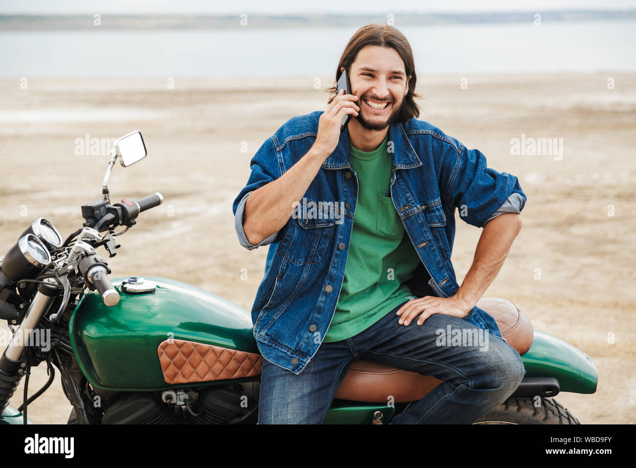 Beau jeune homme portant tenue décontractée assis sur une moto à la plage, talking on mobile phone Banque D'Images
