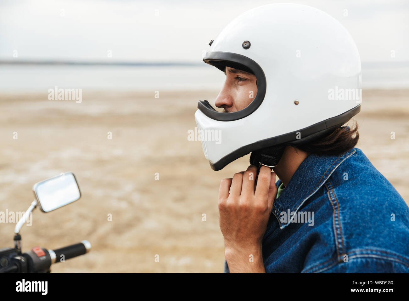 Close up of a young man wearing tenue décontractée assis sur une moto à la plage, mettre sur un casque Banque D'Images