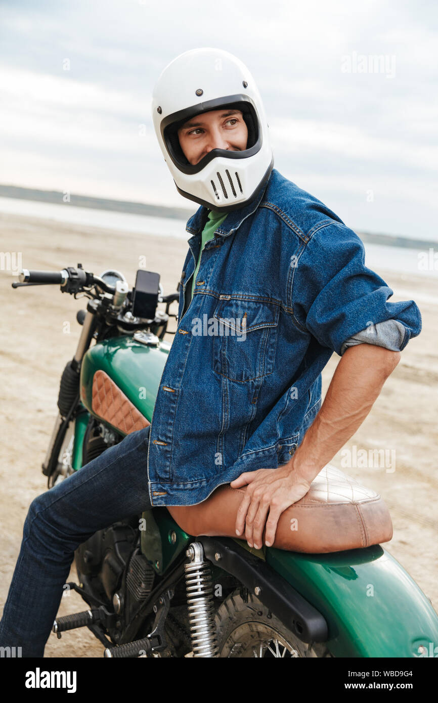 Beau jeune homme portant tenue décontractée assis sur une moto à la plage, le port d'un casque Banque D'Images