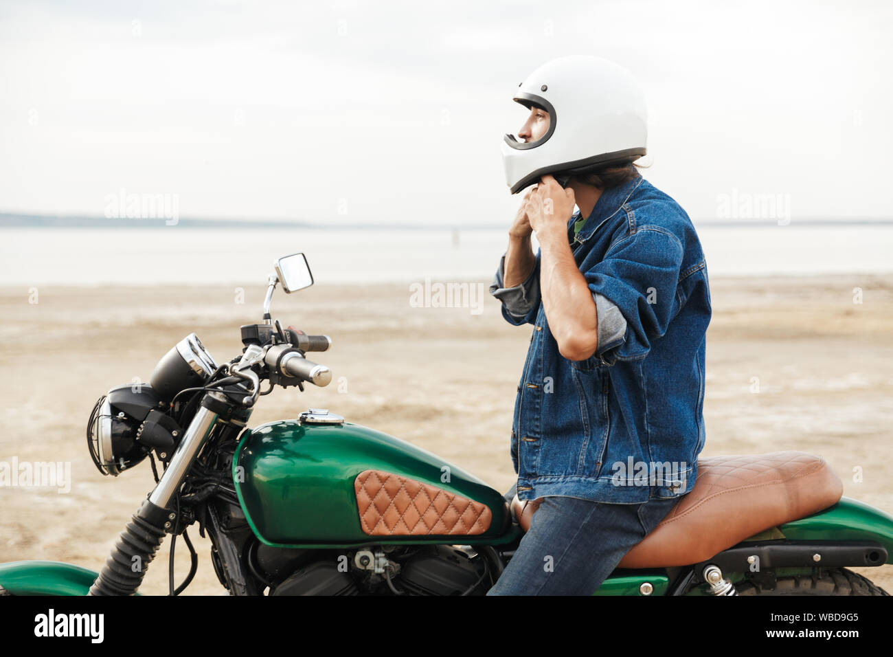 Vue latérale d'un beau jeune homme portant tenue décontractée assis sur une moto à la plage, le port d'un casque Banque D'Images