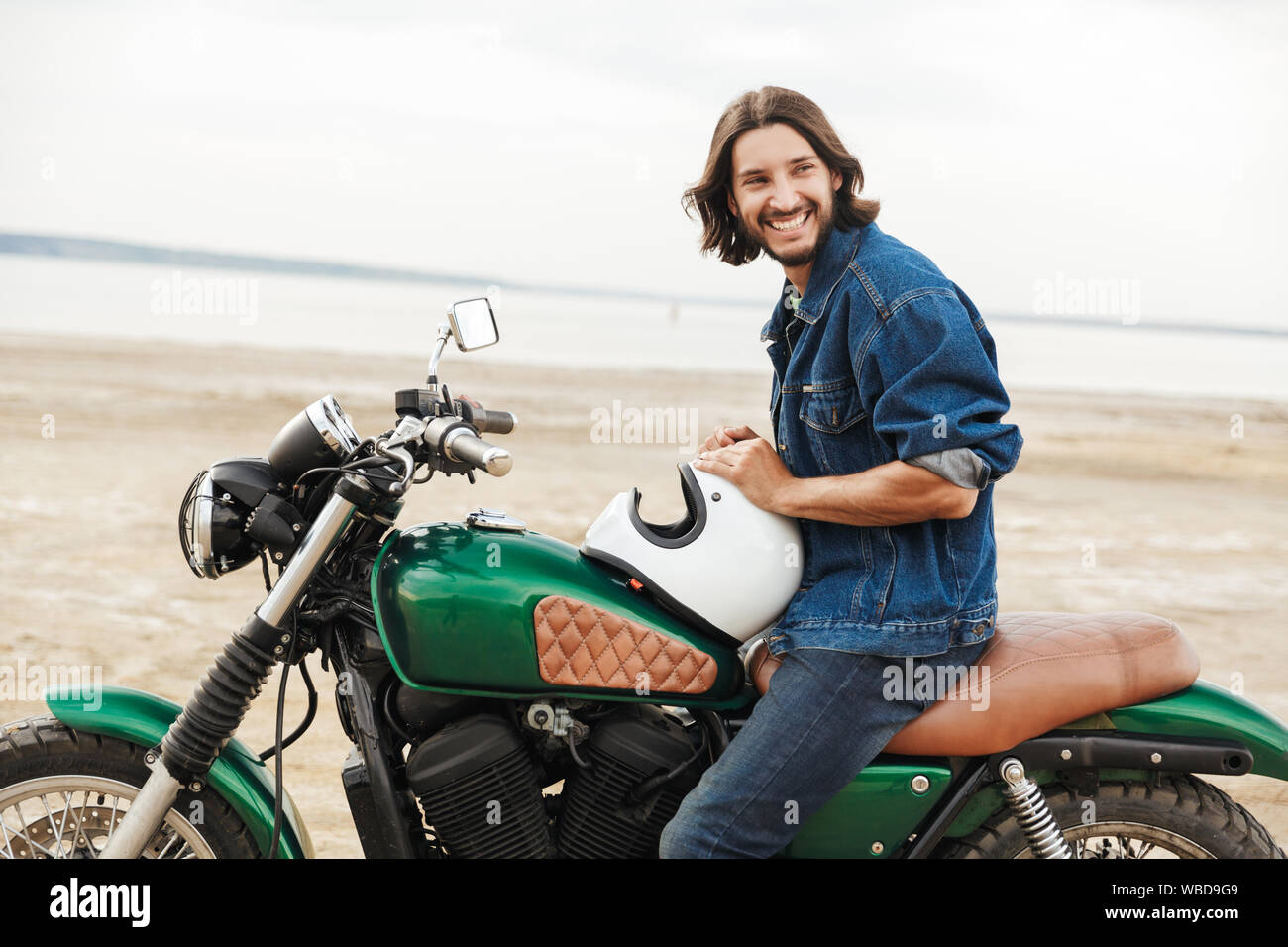 Vue latérale d'un beau jeune homme portant tenue décontractée assis sur une moto à la plage Banque D'Images