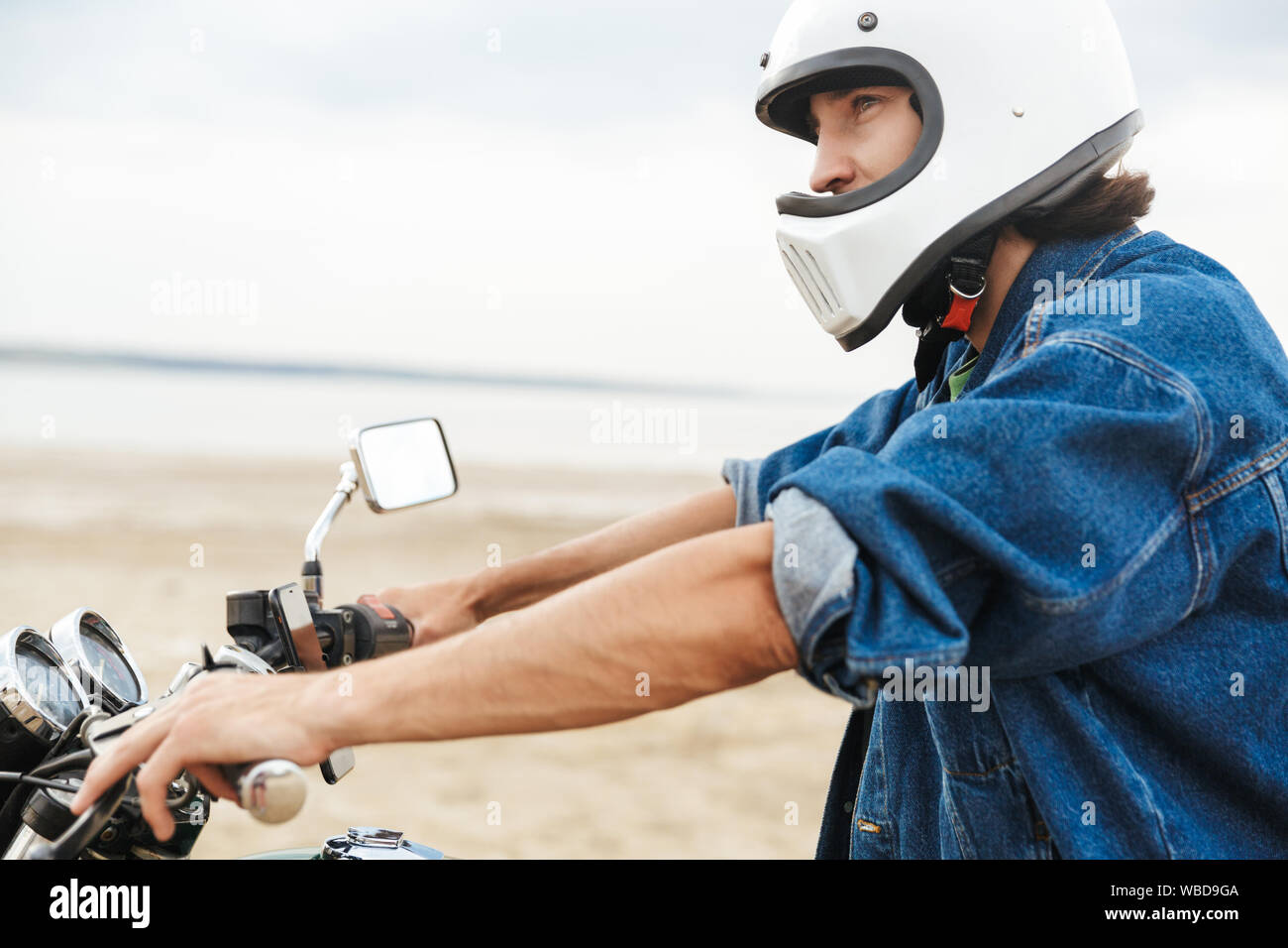 Vue latérale d'un beau jeune homme portant tenue décontractée assis sur une moto à la plage, le port d'un casque Banque D'Images