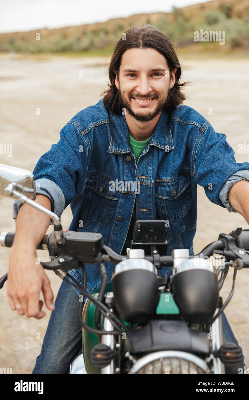 Close up d'un beau jeune homme portant tenue décontractée assis sur une moto à la plage Banque D'Images