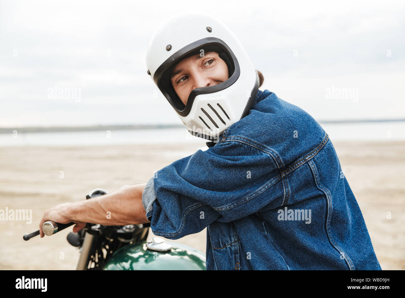 Beau jeune homme portant tenue décontractée assis sur une moto à la plage, le port d'un casque Banque D'Images