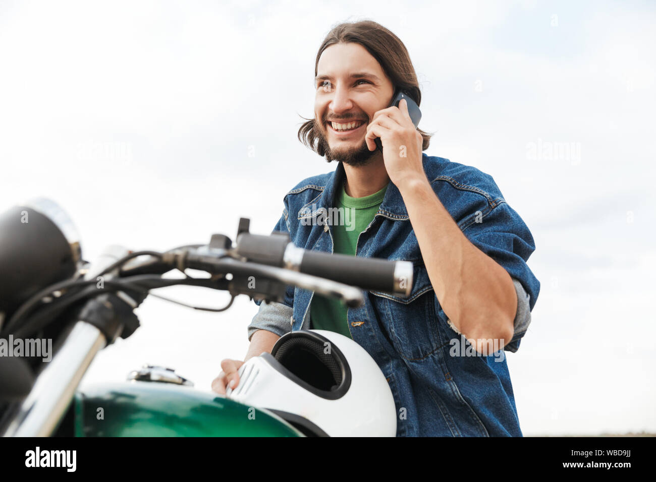 Close up d'un beau jeune homme portant tenue décontractée assis sur une moto à la plage, talking on mobile phonw Banque D'Images