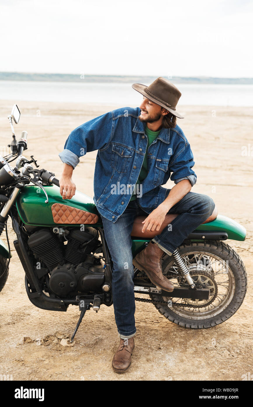 Close up d'un beau jeune homme portant tenue décontractée assis sur une moto à la plage Banque D'Images
