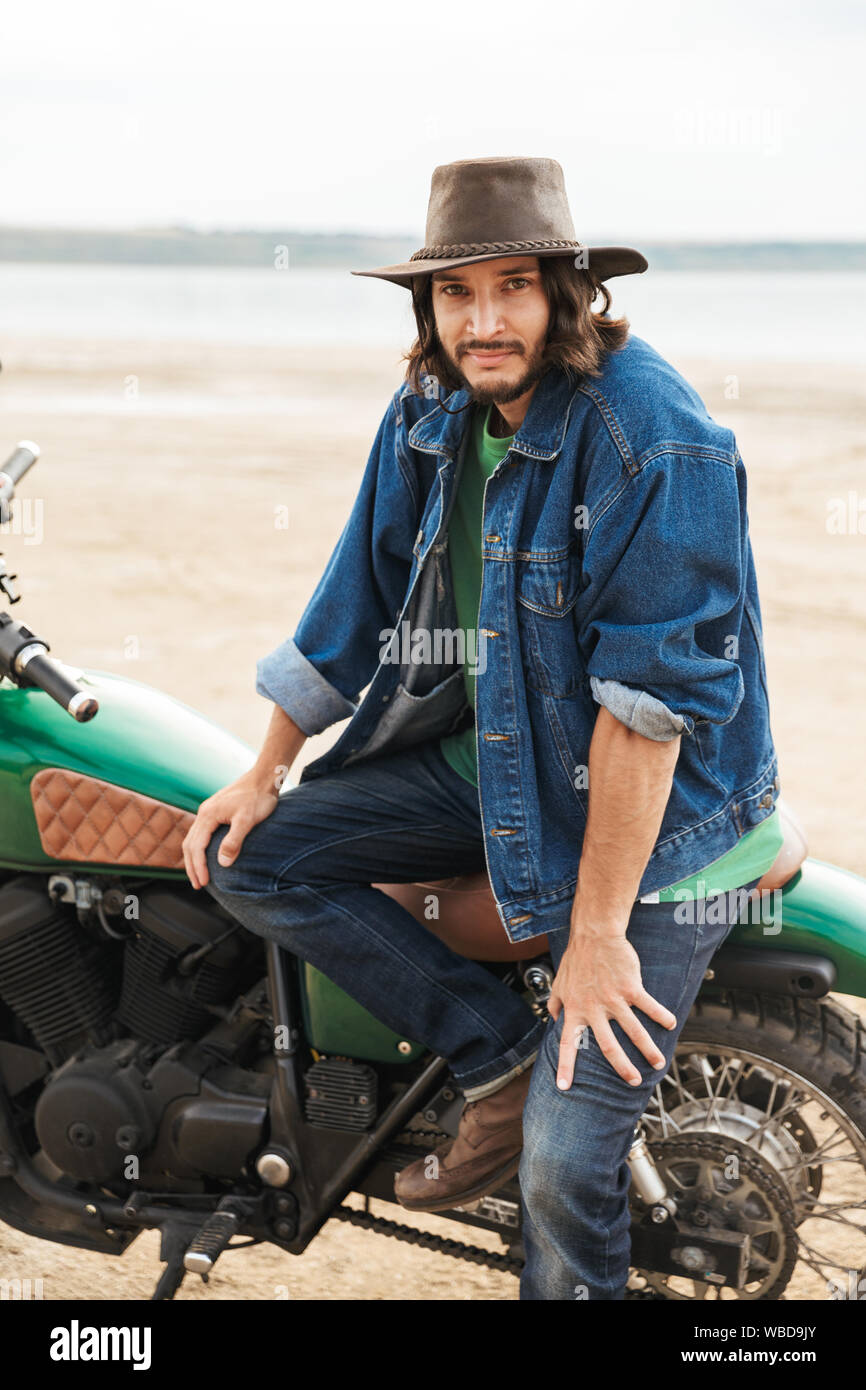 Close up d'un beau jeune homme portant tenue décontractée assis sur une moto à la plage Banque D'Images