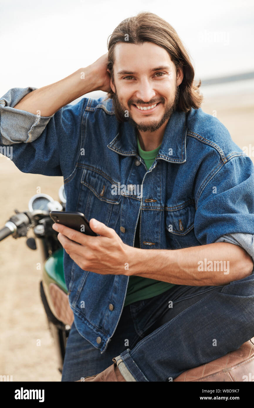 Close up d'un beau jeune homme portant tenue décontractée assis sur une moto à la plage, holding mobile phone Banque D'Images
