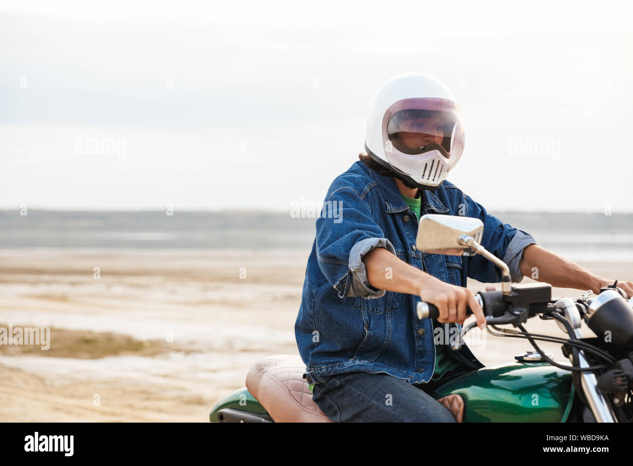 Close up of a young man wearing tenue décontractée assis sur une moto à la plage, le port d'un casque Banque D'Images