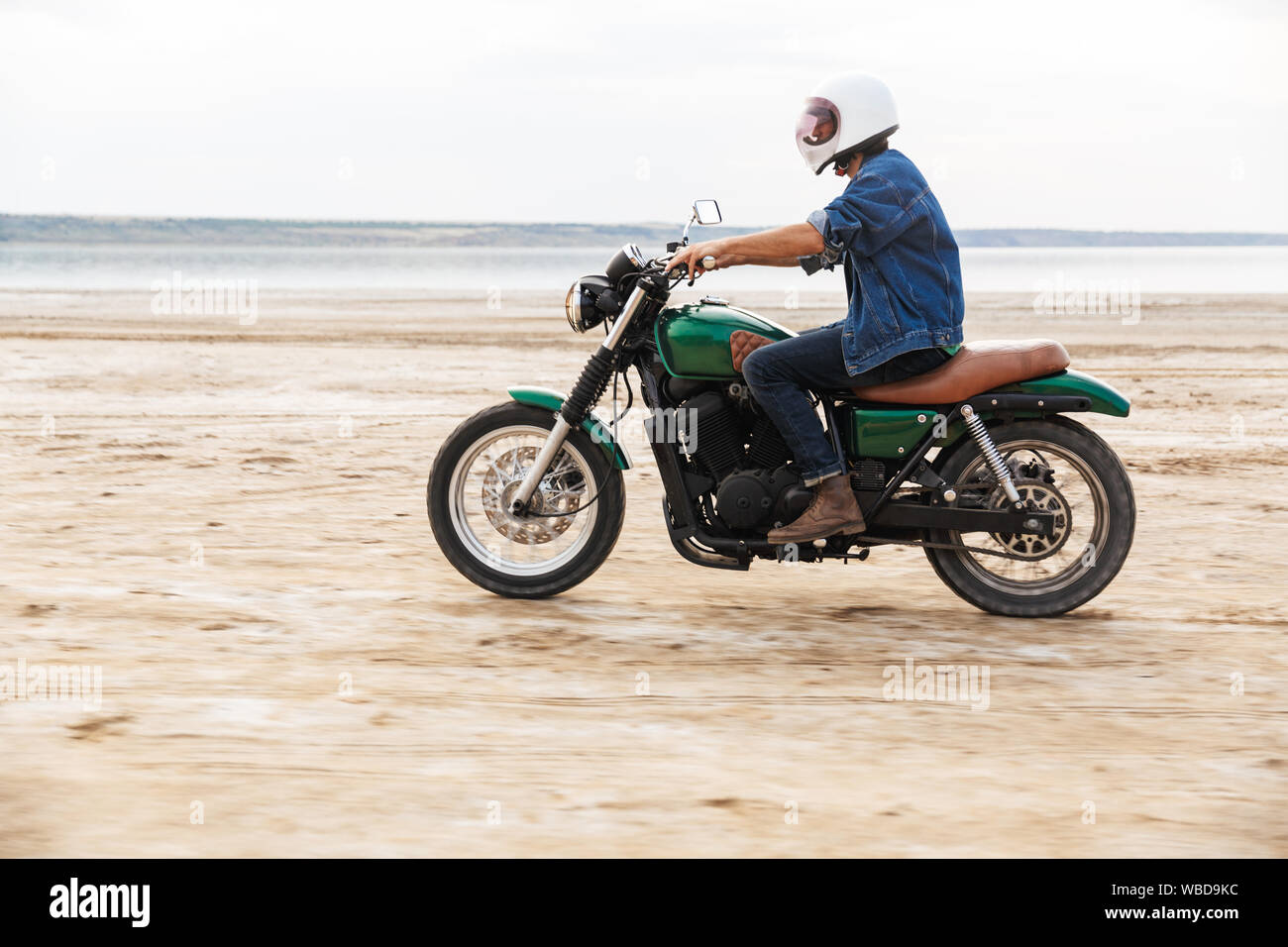 Beau jeune homme portant tenue décontractée circonscription une moto à la plage, le port d'un casque Banque D'Images