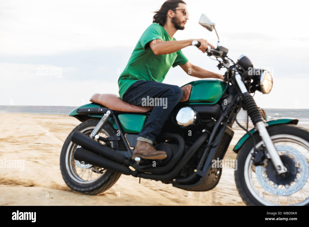 Vue latérale d'un beau jeune homme portant tenue décontractée circonscription une moto à la plage Banque D'Images