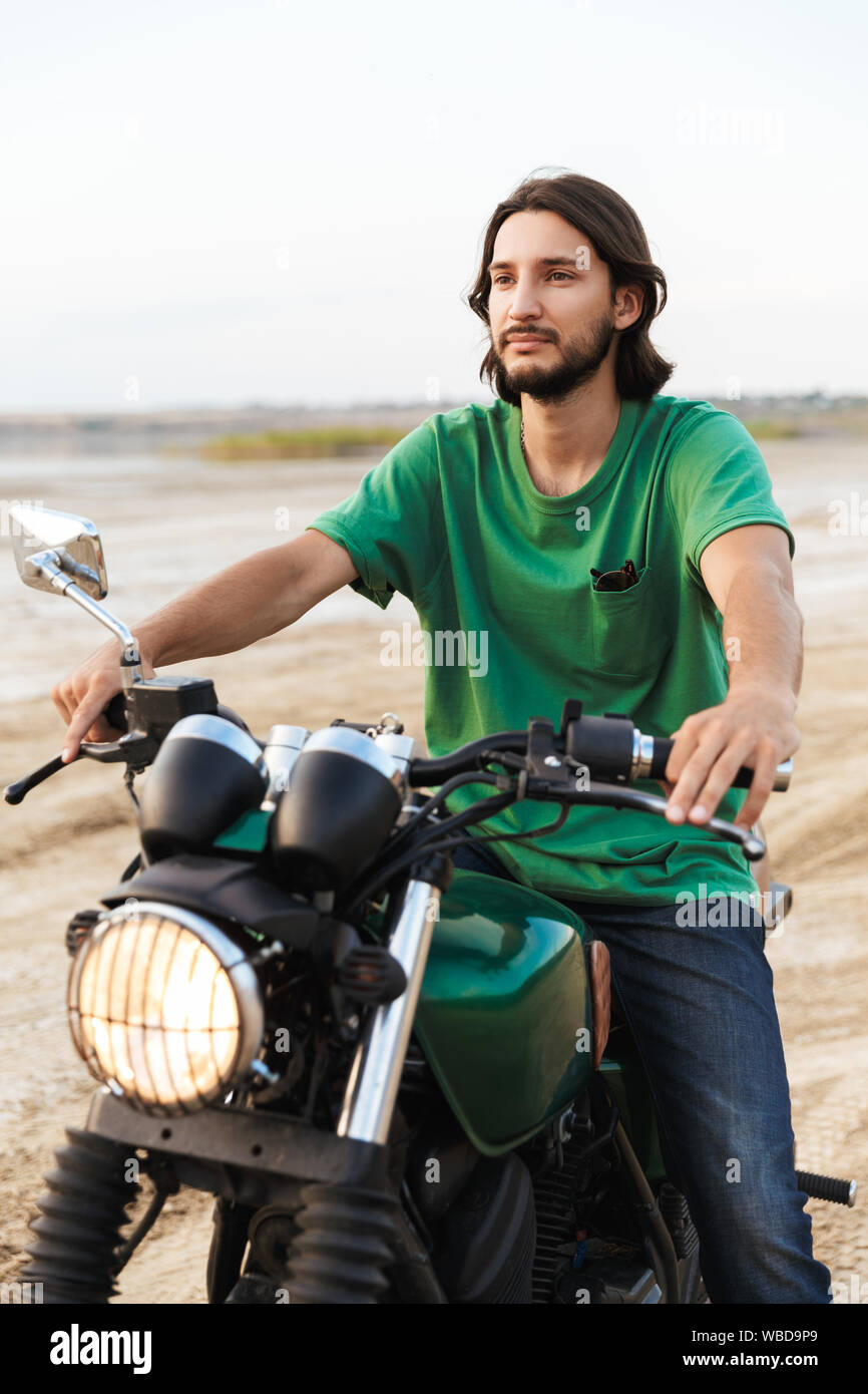 Beau jeune homme portant tenue décontractée assis sur une moto à la plage Banque D'Images