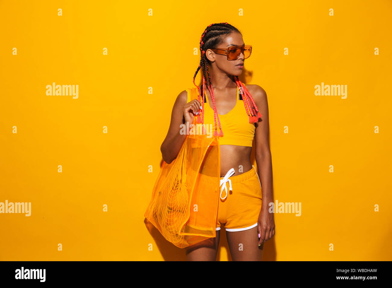 Image of african american woman wearing sunglasses debout et portant le sac en plastique sur fond jaune isolé Banque D'Images