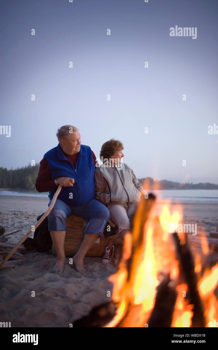 Happy senior couple assis à côté d'un petit feu sur une plage de sable. Banque D'Images