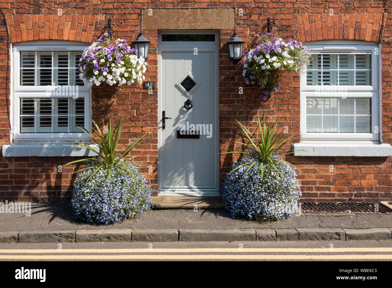 Vieux chalet moderne en brique rouge avec double vitrage PVC et porte avant avec de jolies fleurs en pots et paniers suspendus, Uppingham, Rutland, UK. Banque D'Images