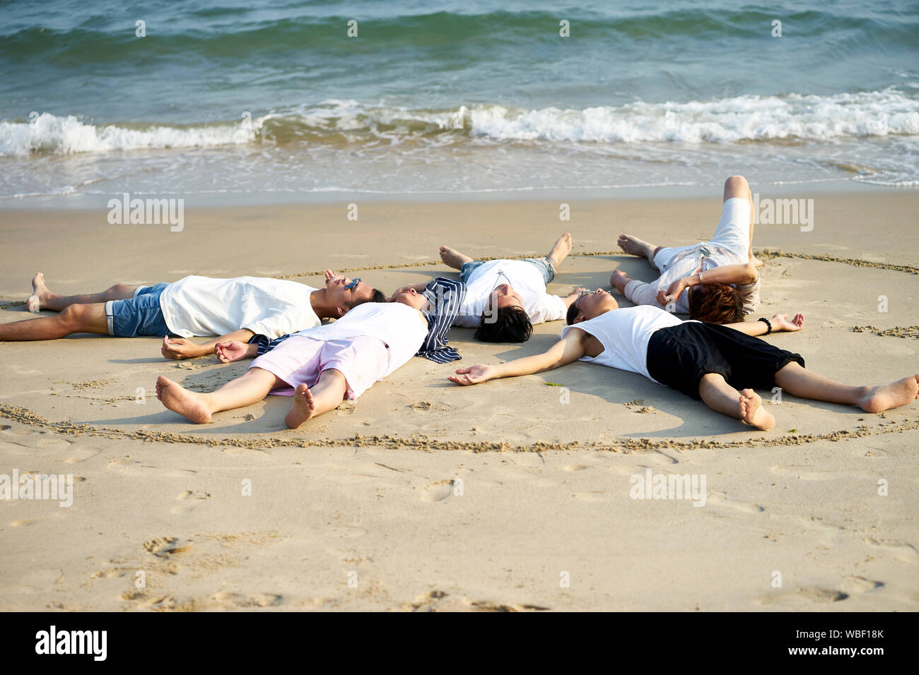 Groupe de cinq jeunes adultes asiatiques hommes couchés détendu sur plage de sable fin formant un cercle Banque D'Images