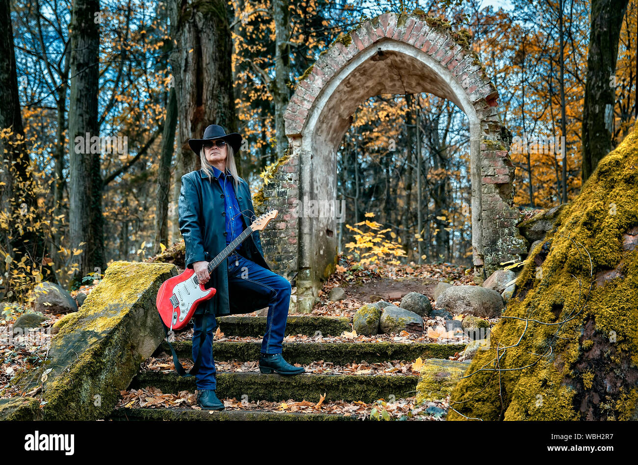 Personnes âgées l'homme aux cheveux longs dans un manteau de cuir, chapeau, lunettes de soleil et un jean debout avec une guitare électrique dans une sombre forêt près d'une chapelle en ruine Banque D'Images