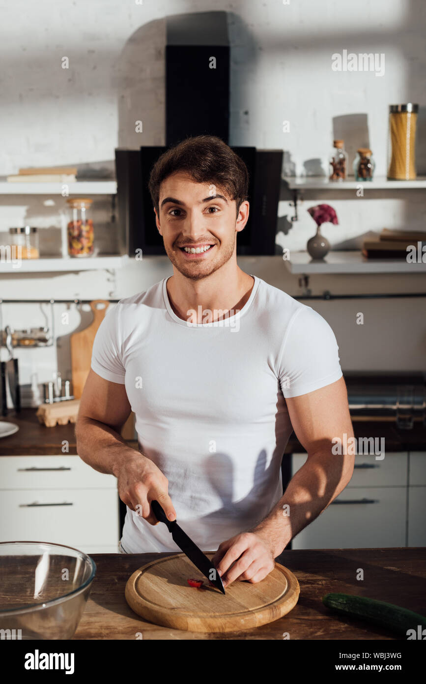 Homme musclé en souriant blanc T-shirt couper des légumes dans la cuisine Banque D'Images