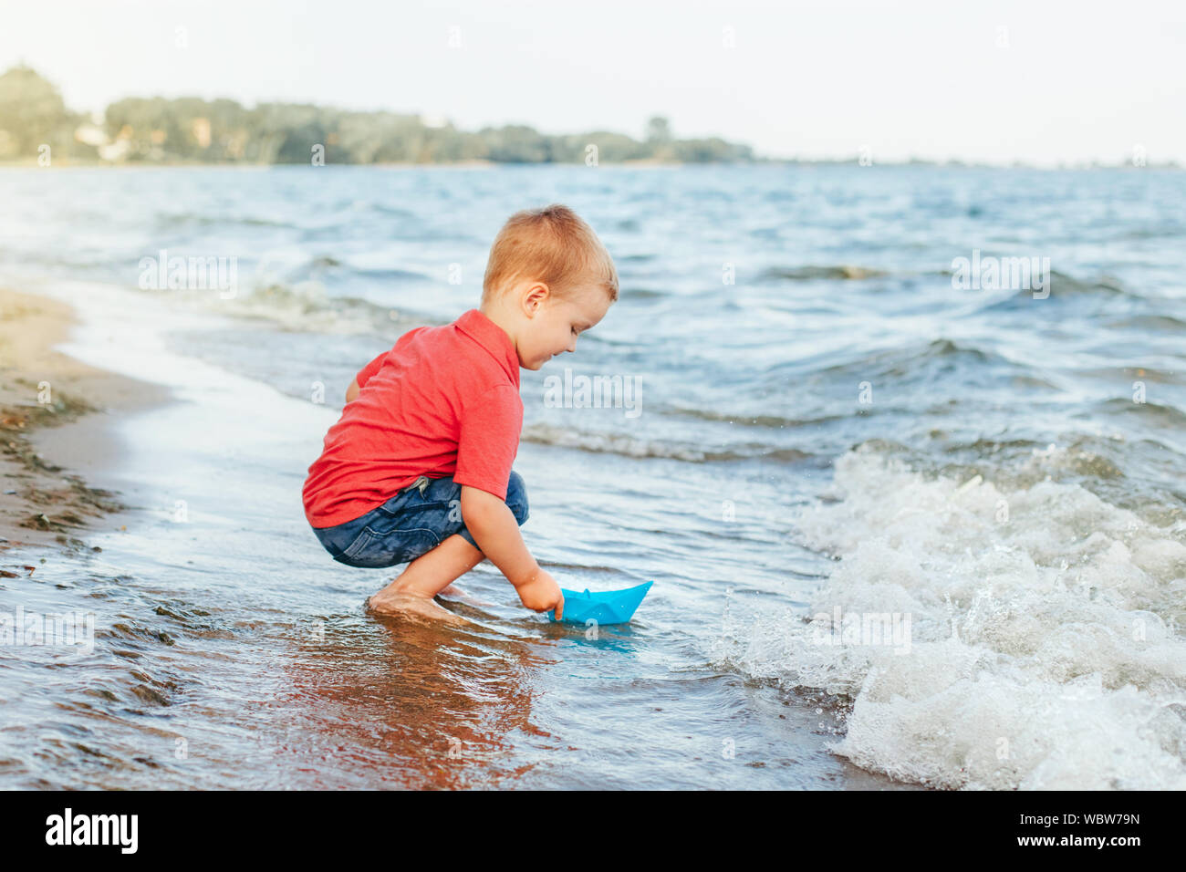 Heureux rousse caucasien enfant bébé garçon Mettre papier bleu voile dans  l'eau sur le lac mer océan port au coucher du soleil du soir ou matin.  Enfant jouant sur bea Photo Stock -