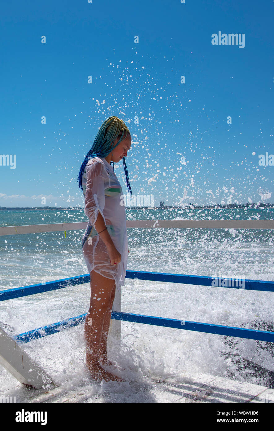 Une fille mince en maillot blanc et bleu, tunique avec tresses sénégalaises,  se dresse sur un balcon au-dessus de la mer. Briser les vagues sur des  pierres et splatter scatt Photo Stock -