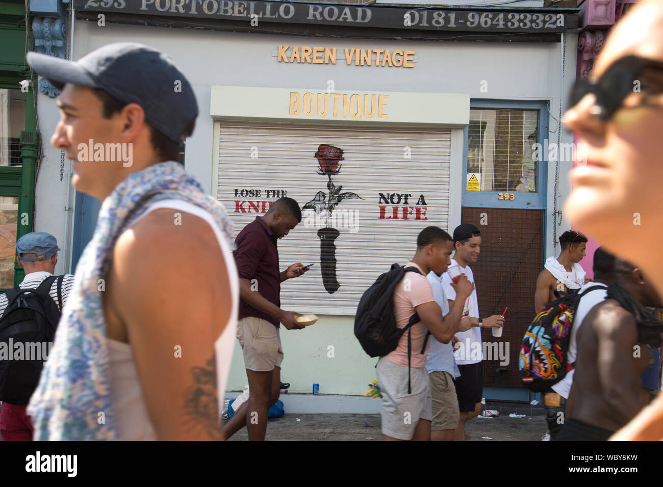 Le carnaval de Notting Hill, le 26 août 2019. Les jeunes passent devant une boutique avec le slogan "Perdre le couteau, pas une vie" peint sur son obturateur. Banque D'Images