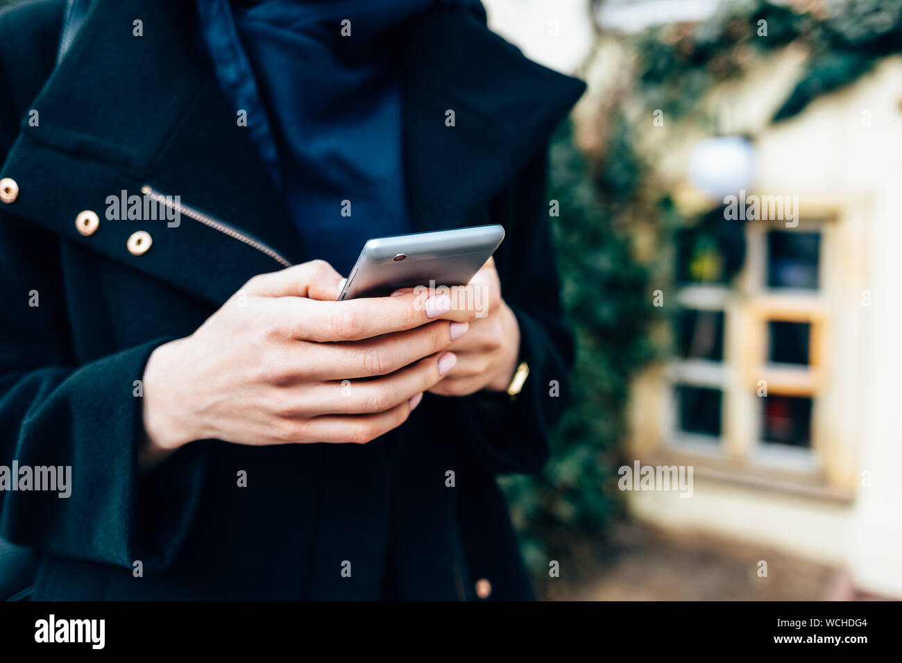 Jeune femme vêtue d'un élégant manteau noir holding mobile phone debout sur la rue de ville en journée, close-up. Banque D'Images