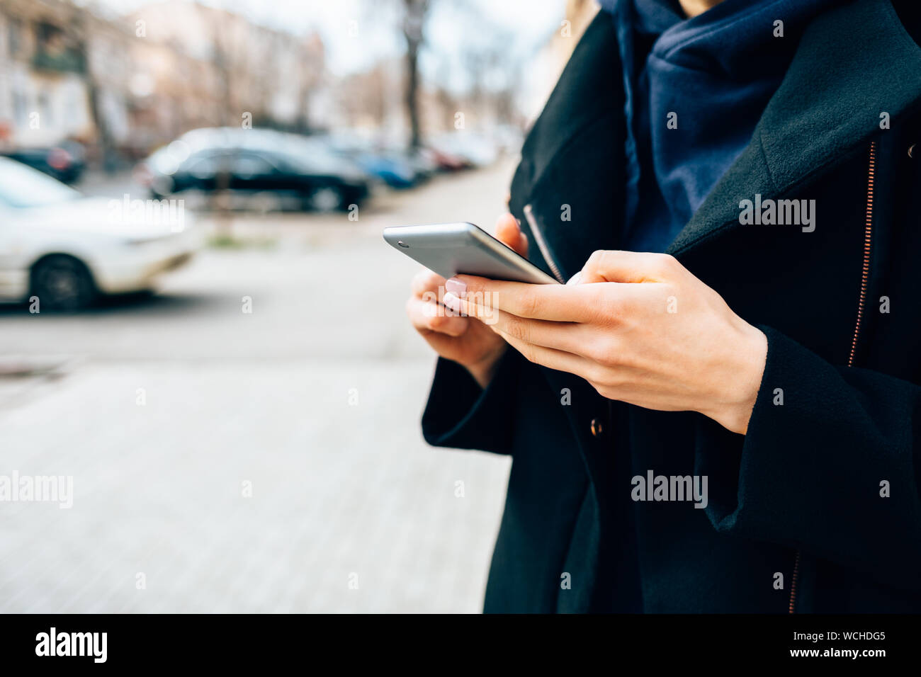 Close-up jeune femme portant un élégant manteau noir holding mobile phone debout sur la rue de ville près de la route et des voitures dans la journée. Banque D'Images