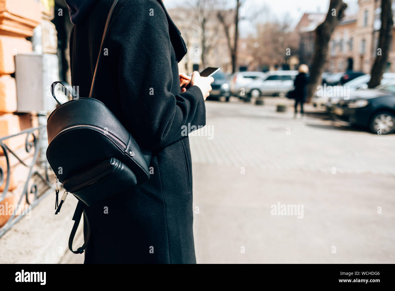 Vue de côté jeune femme portant un sac à dos élégant manteau noir et à l'aide de mobile phone debout sur street dans la vieille ville européenne près de road avec des voitures dans daytim Banque D'Images