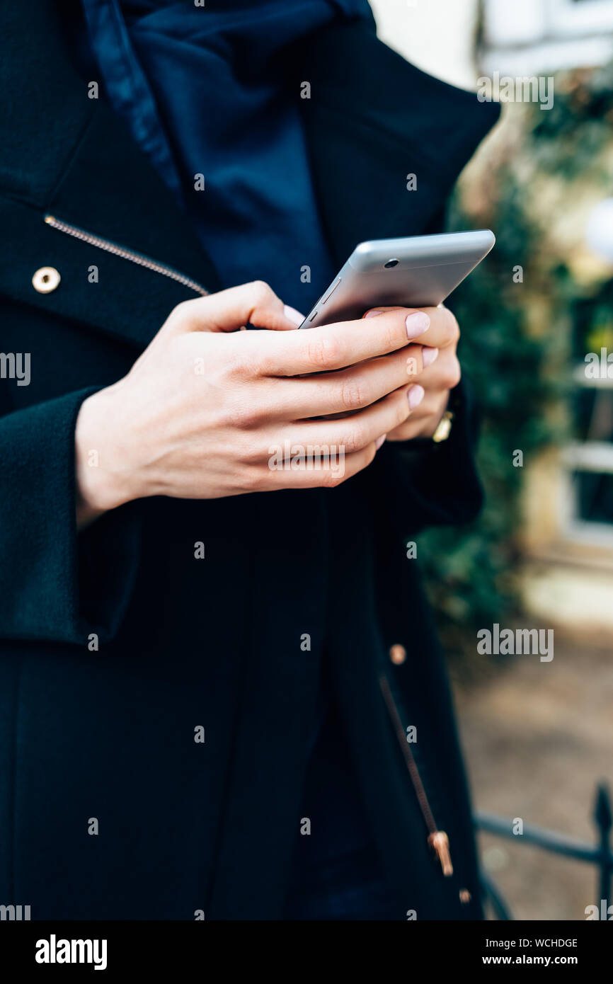 Close-up jeune femme vêtue d'un élégant manteau noir tenant debout sur téléphone mobile de rue ville de jour. Banque D'Images