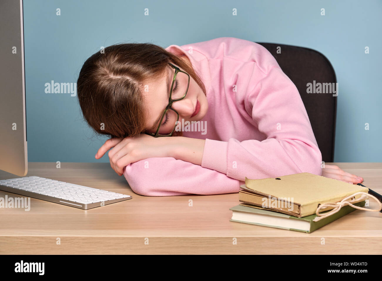 Une fille dans un chandail rose est assis à une table avec un ordinateur et des livres avec sa tête reposant sur les mains. Banque D'Images