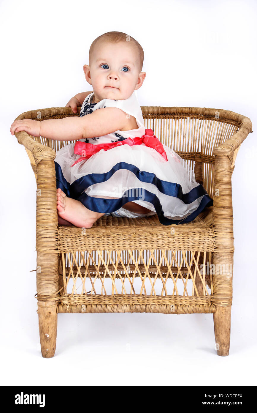 6 mois bébé fille dans une jolie robe assis sur une chaise en rotin Banque D'Images