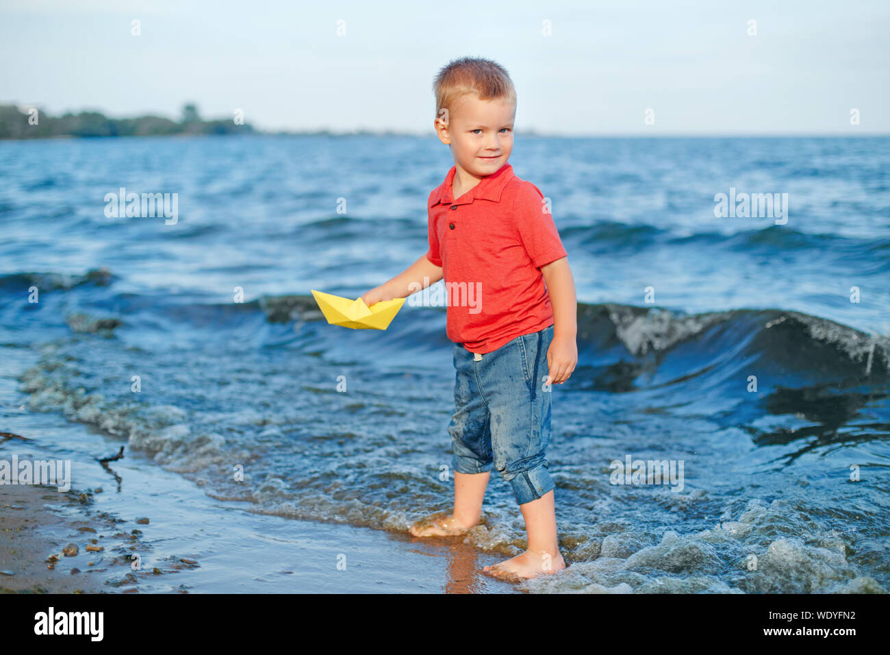 Heureux rousse caucasien enfant bébé garçon Mettre papier jaune en bateau  sur le lac de l'eau mer océan port au coucher du soleil du soir ou matin.  Enfant jouant sur b Photo