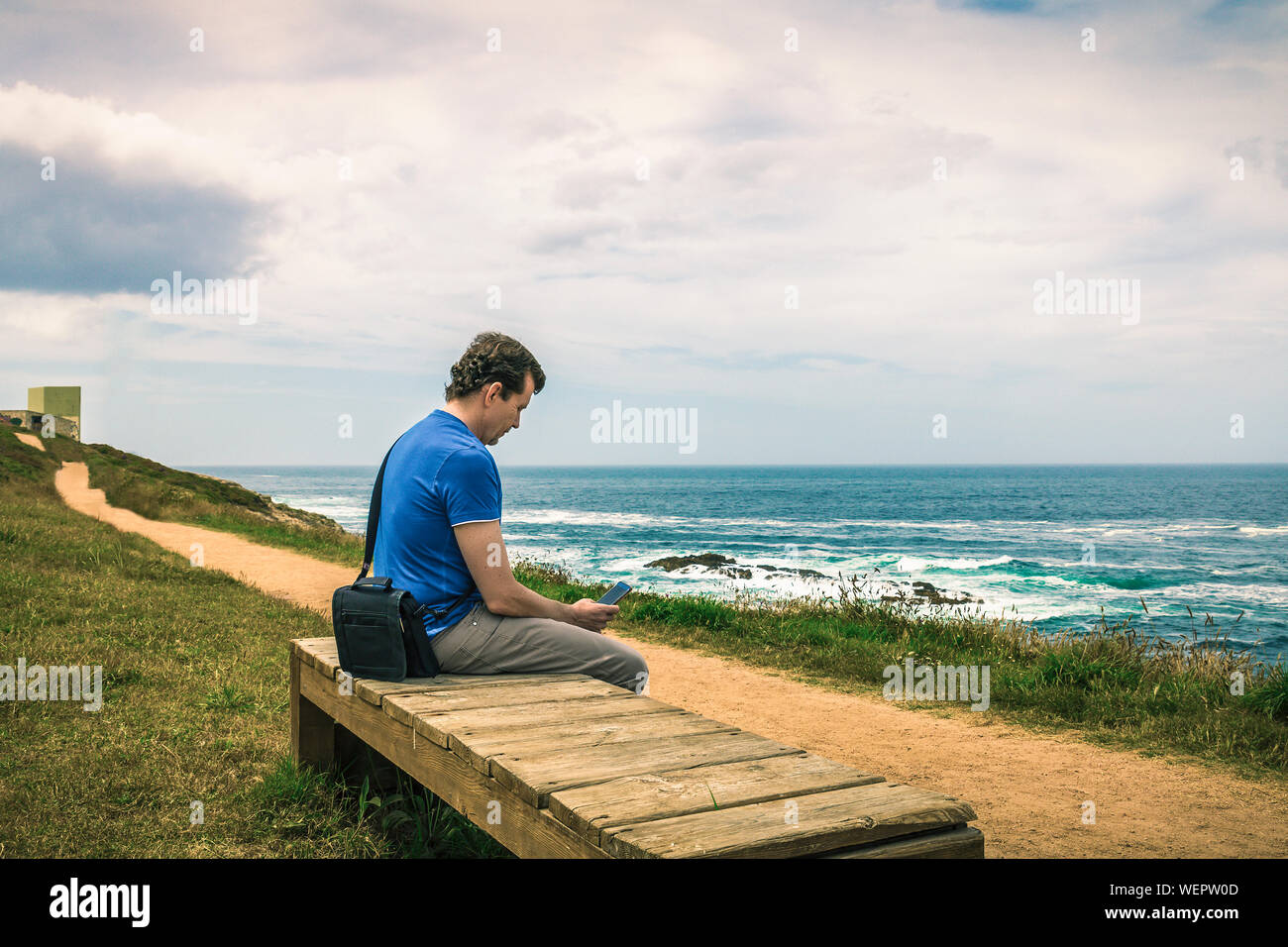 Homme avec chemise bleue assis sur un banc en bois en regardant son téléphone intelligent face à la mer Banque D'Images