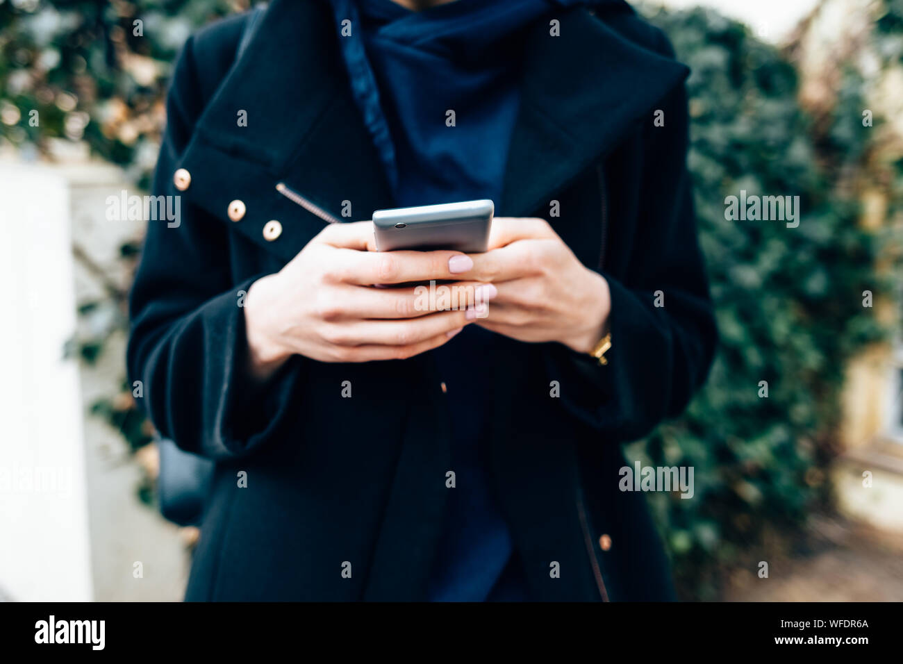 Jeune femme portant un élégant manteau noir holding mobile phone standing outdoors dans journée, close-up. Banque D'Images