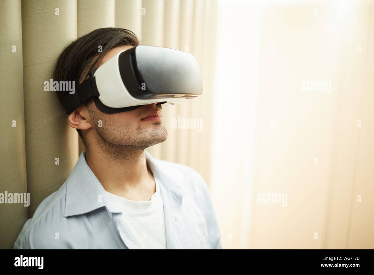 Head and shoulders portrait d'un homme portant un casque VR leaning against wall en salle vide, copy space Banque D'Images