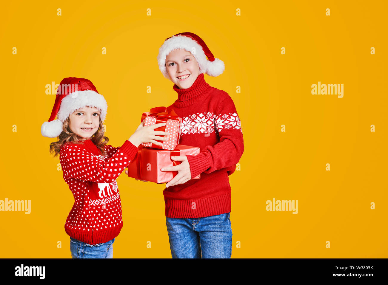 Happy kids in Santa hats et chandails rouge avec permanent présente à rire et à la recherche à l'appareil photo sur fond jaune Banque D'Images