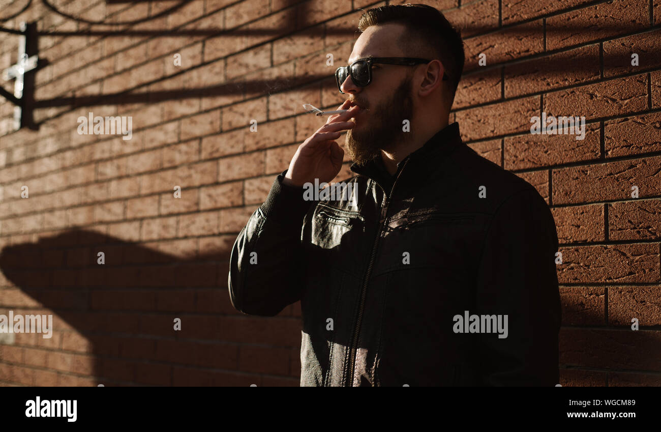 Vue latérale du beau barbu fumer une cigarette sur mur de brique Banque D'Images