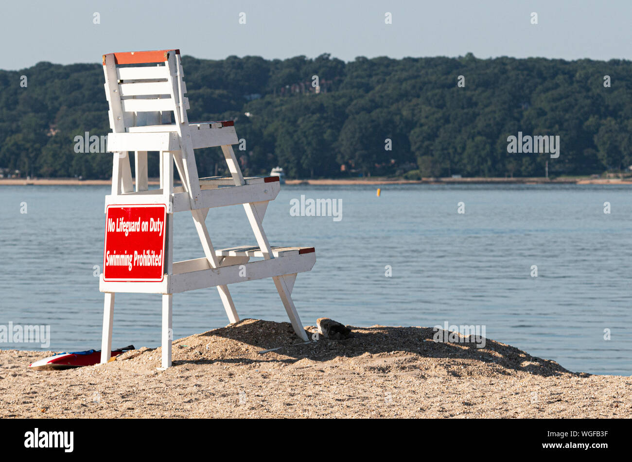 Un sauveteur blanc debout sur le sable à la recherche sur l'eau vide de la rive nord de Long Island, avec des collines et des arbres en arrière-plan. Banque D'Images