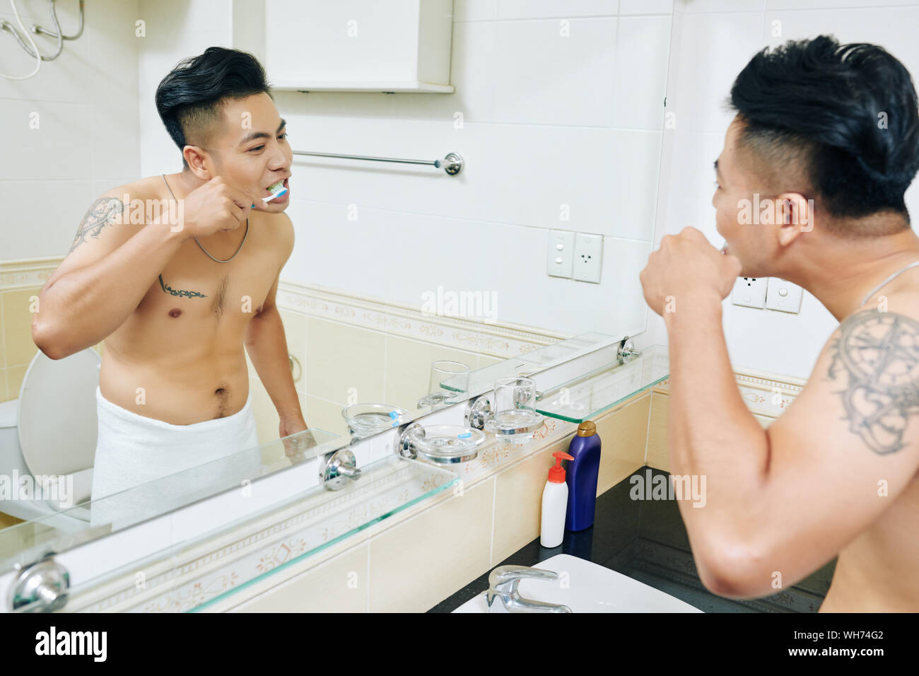 Jeune homme vietnamien enveloppé dans une serviette après la douche se brosser les dents en face de miroir dans salle de bains Banque D'Images