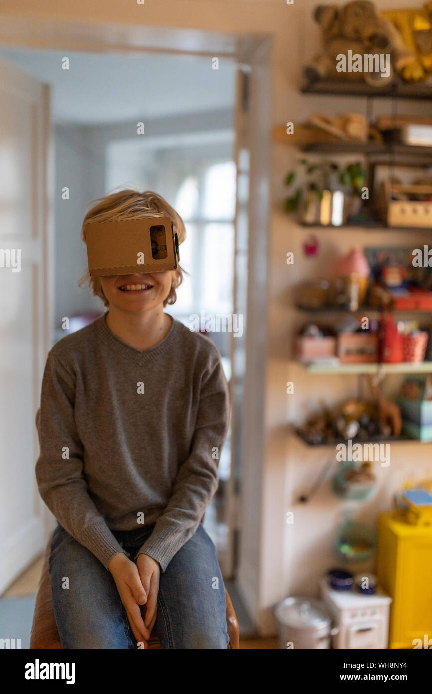 Garçon assis sur l'argent dans sa chambre à la maison portant des lunettes VR en carton Banque D'Images