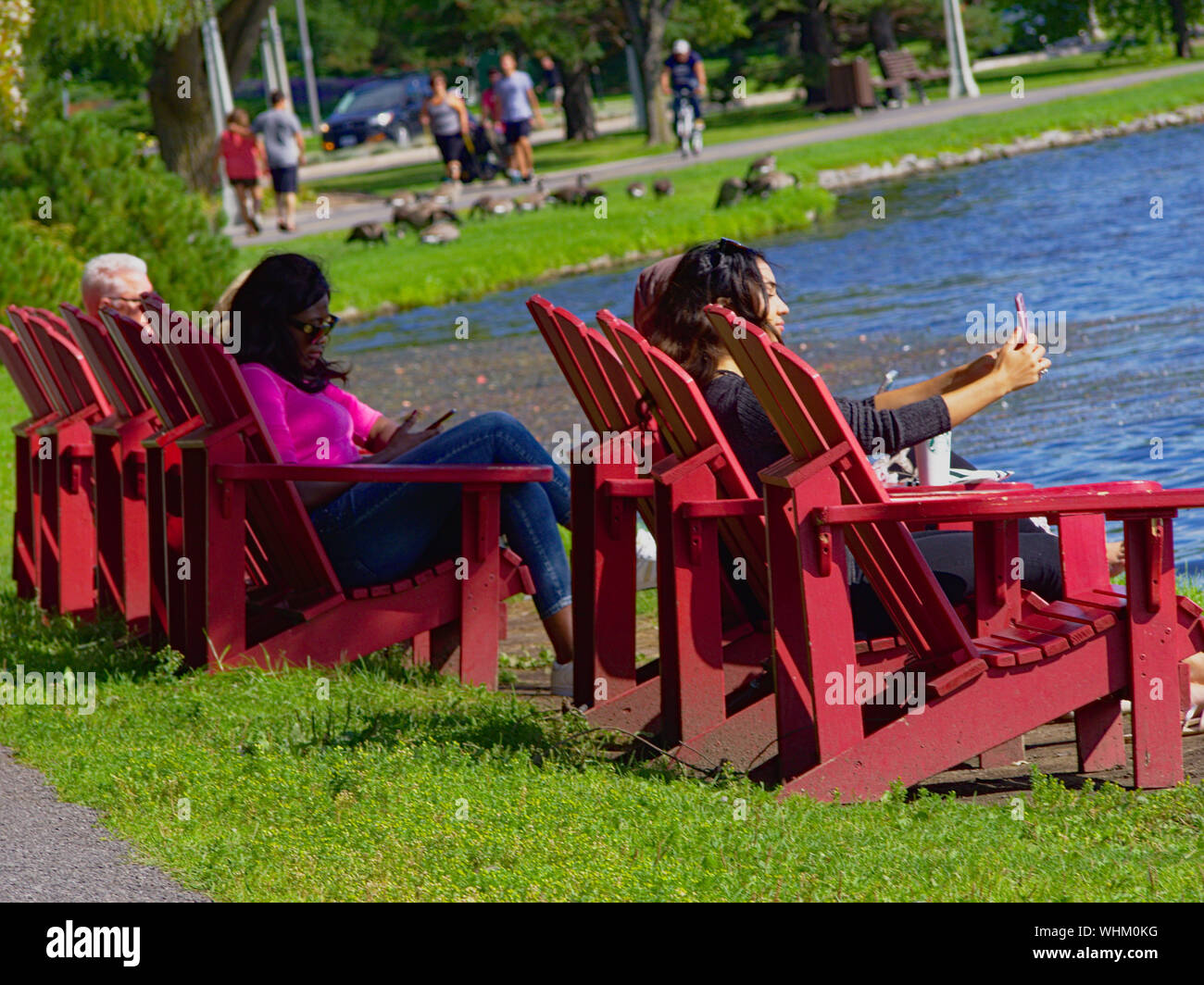 Une jeune femme assise sur une chaise longue rouge au bord d'un lac, prend une de ses selfies et un ami. Ottawa, Ontario, Canada. Banque D'Images