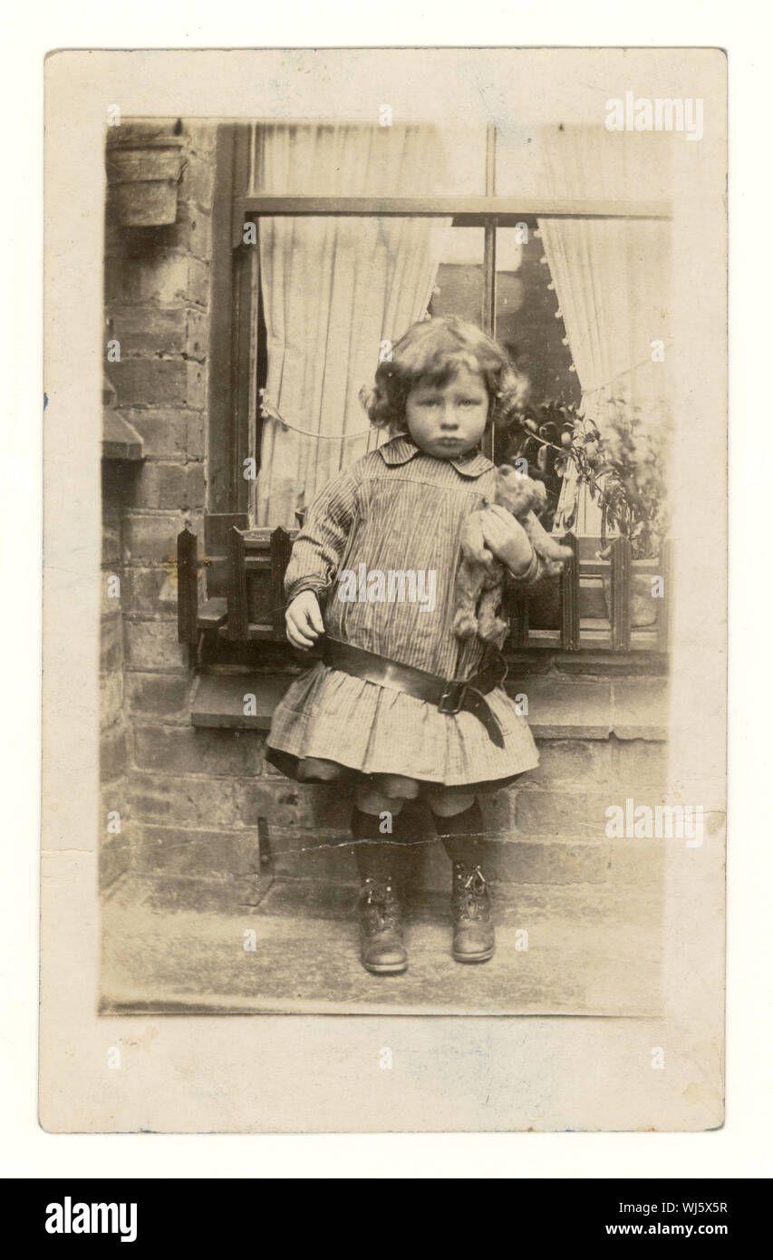 Début des années 1900 Carte postale de jeune enfant tenant un ours en peluche à l'extérieur de maison, avec des robe taille basse avec un ours en peluche, à l'extérieur de maison, avec des robe taille basse avec ceinture, vers 1907, au Royaume-Uni. Banque D'Images