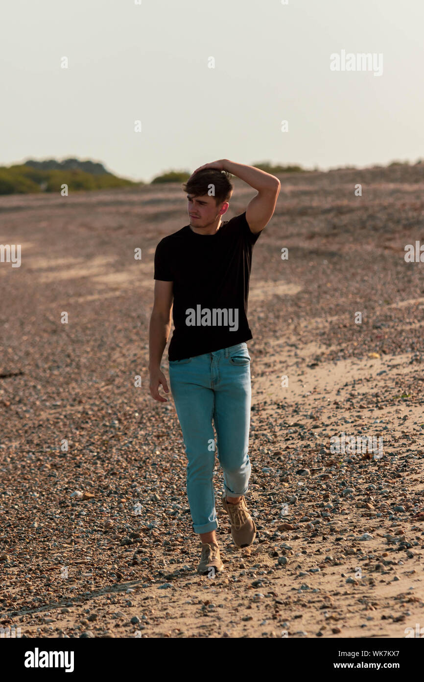 Young caucasian man walking sur une plage à l'heure d'or portant un t-shirt et jeans Banque D'Images