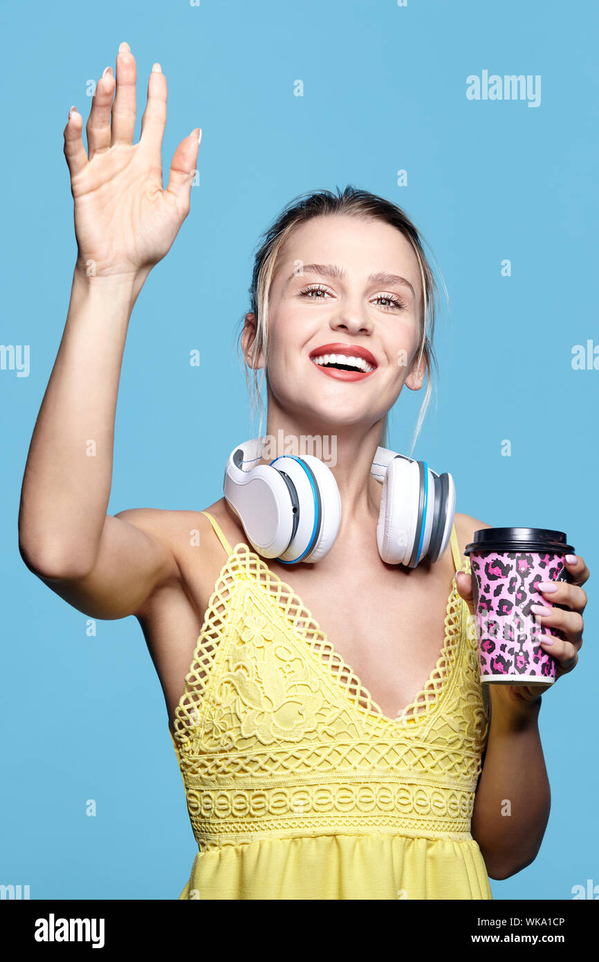 Jolie jeune femme heureuse de boire du café et à écouter de la musique dans le casque. Femme en robe jaune en agitant la main sur fond bleu. Smiling girl wit Banque D'Images