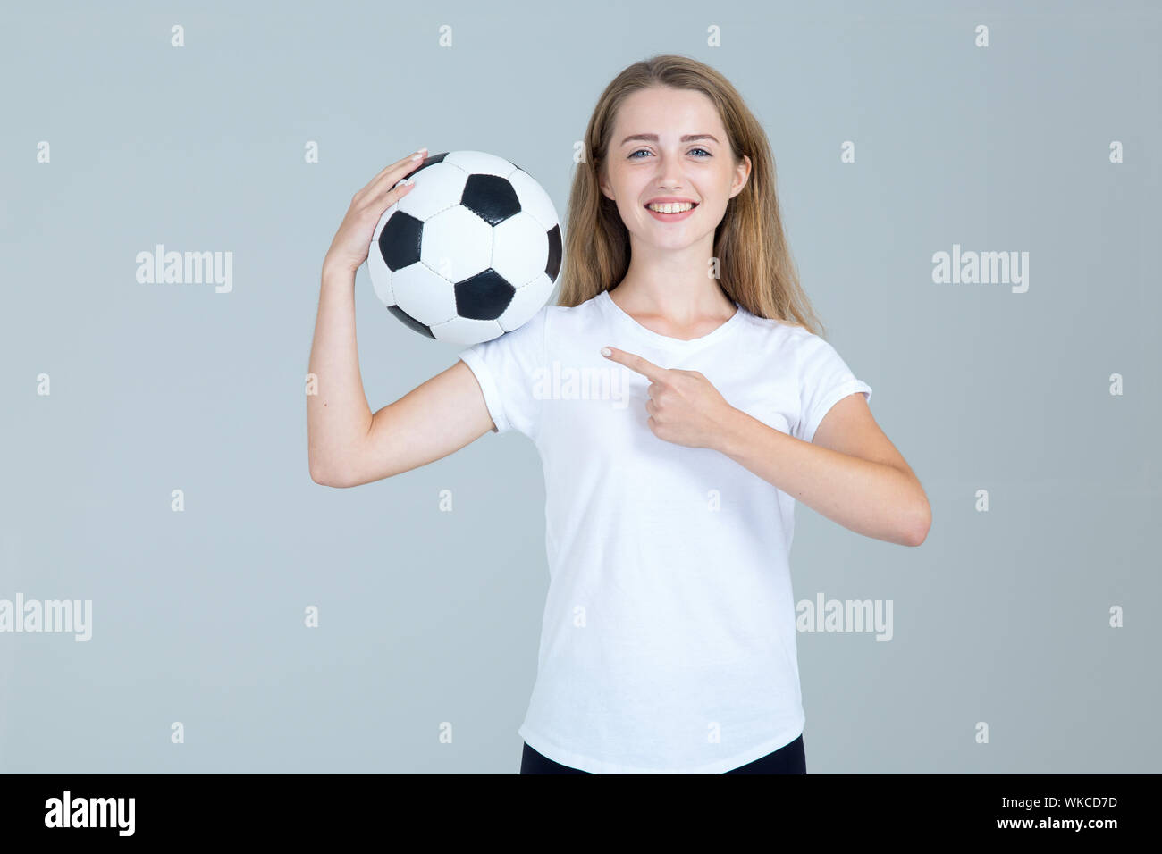 Jeune femme heureuse avec un ballon de football dans ses mains pointe vers la côte sur un fond gris. Fan de football. Banque D'Images